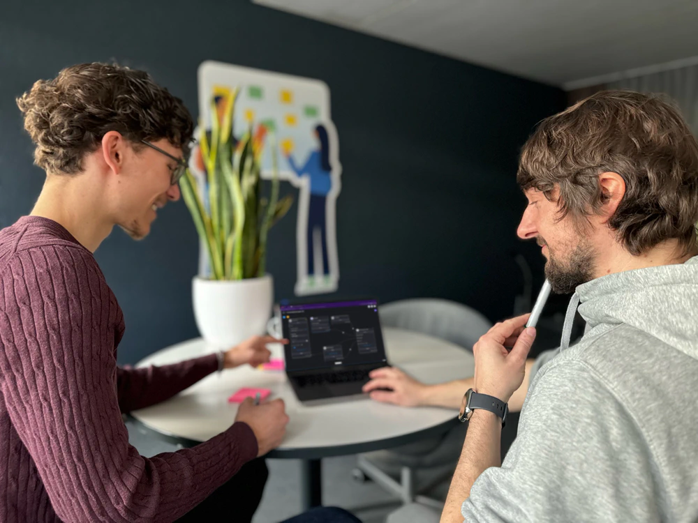 Zwei Personen schauen zusammen auf einen Laptop-Bildschirm. Der Bildschirm zeigt ein zusammenhängende Komponenten eines Modells. Die Person auf der linken Seite zeigt auf den Bildschirm, die Person rechts sieht interessiert zu. Im Hintergrund steht eine Pflanze.